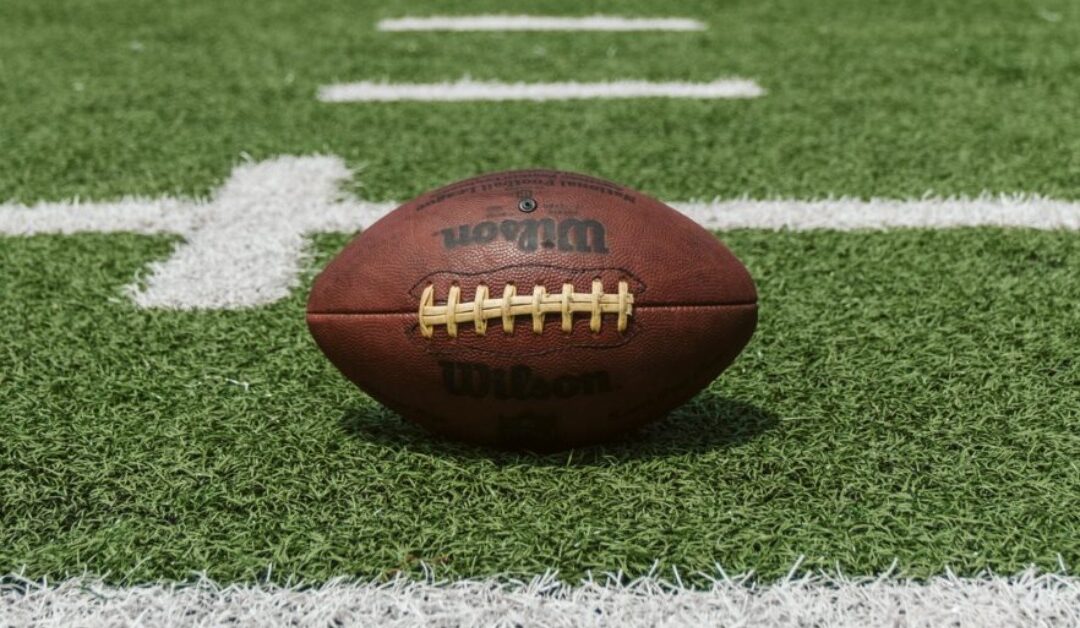 A used-looking football sits on green grass between yard lines on a football field.