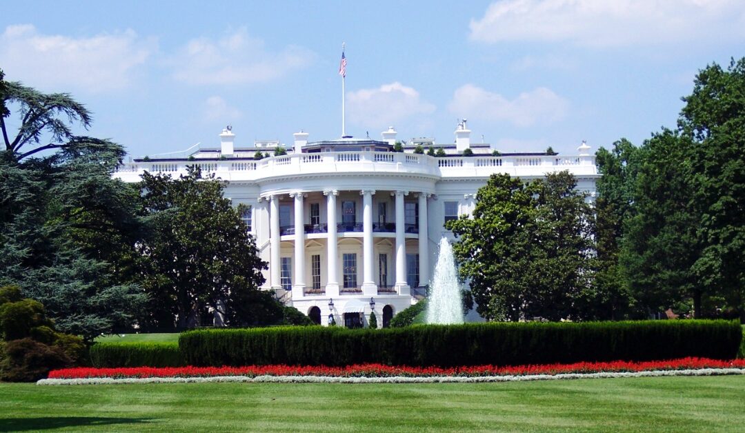 The White House sits behind green grass, shrubs, and trees on a sunny day.