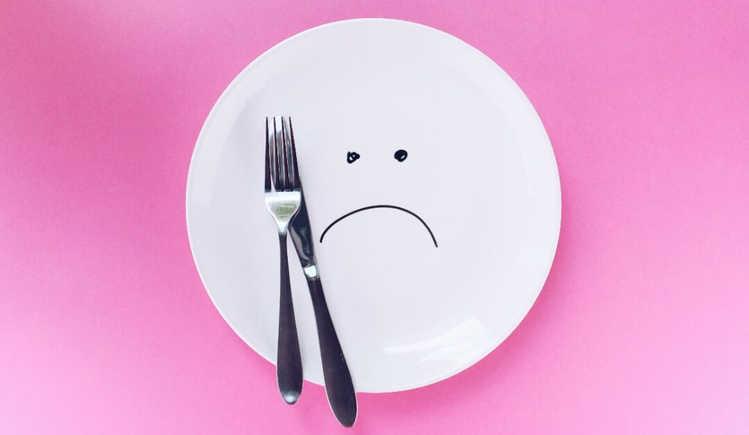 An empty white plate frowns as a fork and knife rest atop it on a pink background.