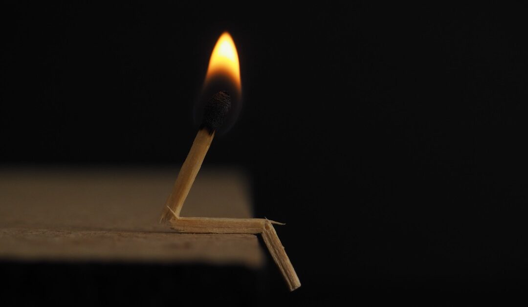 A lit match is bent to resemble a person sitting on the edge of a table, the fire slowly burning out in the dark.