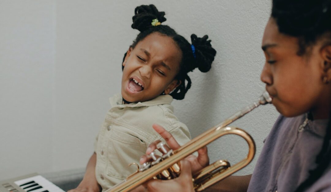 A young girl belts out a song next to an older girl playing a horn.